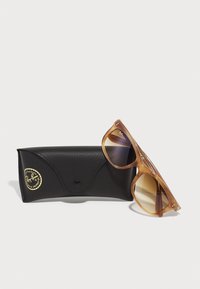 Ochelari de soare Ray-Ban cu o ramă transparentă de culoare maro deschis și lentile degradate maro, alături de o cutie din piele neagră cu un logo auriu.
