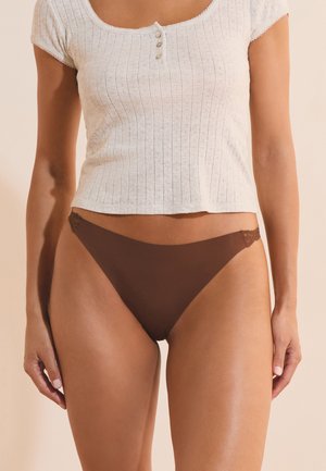 Mujer con ropa interior estilo bikini marrón con detalles de encaje y una camiseta blanca de manga corta acanalada con botones, de pie frente a un fondo neutro.