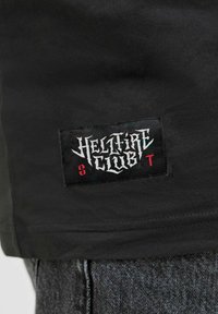 Černé tričko s texturovanou černou nášivkou s nápisem "HELLFIRE CLUB" v stylizovaném bílém a červeném písmu, bavlněný materiál, krátké rukávy.