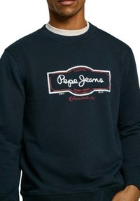 Felpa blu navy in cotone con scollatura rotonda. Caratterizzata da un prominente logo e testo "Pepe Jeans" in bianco e rosso. Maniche lunghe con polsini a costine.