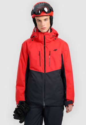 Male skier wearing red and black jacket and helmet, holding black gloves in right hand, standing against plain light background.