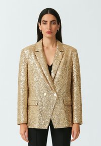 Blazer in paillettes dorate con un design a doppio petto, rever e tasche frontali. Texture liscia con una finitura lucente.