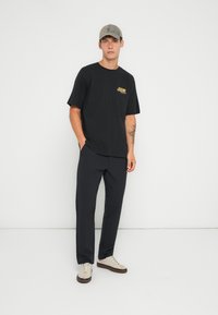 Zwarte T-shirt met korte mouwen en grafische logo, gecombineerd met donkere broeken. Beige sneakers hebben een bruine zool. Grijze pet voegt een casual tintje toe.