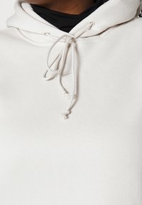 Světlý béžový mikina s kapucí na stažení, jemná textura a minimalistický design, na přední straně s malým bílým logem.