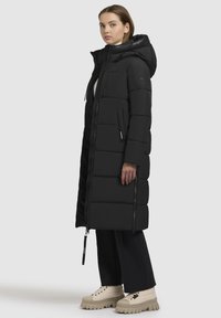 Piumino nero con cappuccio, cuciture verticali e tasche con cerniera. Motivo testurizzato con imbottitura spessa, indossato sopra pantaloni neri e stivali beige.