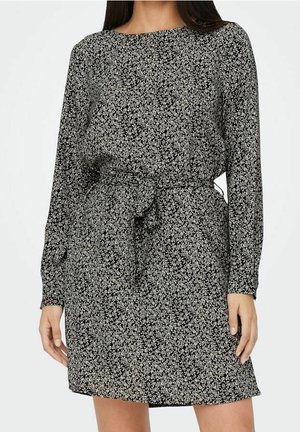 Frau trägt ein schwarz-weiß gemustertes, langärmeliges Kleid mit Blumenmuster und einem gebundenen Taillengürtel, steht vor einem einfarbigen Hintergrund.