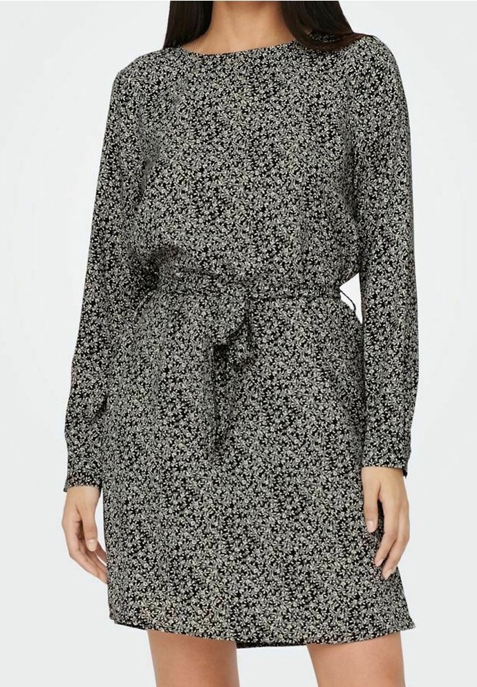 Femme portant une robe longue à manches longues à motif floral noir et blanc avec une ceinture nouée à la taille, se tenant devant un fond uni.