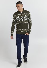 Pull en maille vert foncé avec des motifs géométriques blancs. Dispose d'un col zippé et de poignets côtelés, associé à un jean bleu foncé et des baskets noires.