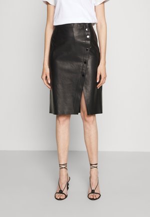 Femme portant une jupe en cuir noire boutonnée avec une fente à l'avant, associée à des sandales noires à talons hauts et à brides, et un haut blanc à manches courtes.