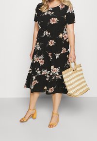 Musta kukallinen mekko lyhyillä hihoilla, jossa on virtaviivainen hame ja laskoksia. Yhdistettynä kudottuun raidalliseen laukkuun ja keltaisiin korkokenkiin.