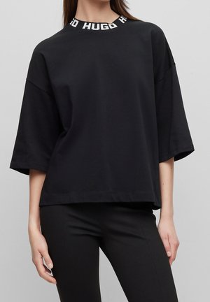 Femme portant une chemise noire ample à manches courtes avec un col montant blanc inscrit "HUGO", associée à un pantalon noir ajusté, debout devant un mur gris uni.