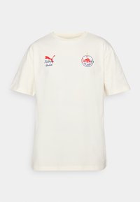 Krem majica od pamuka s kratkim rukavima. Sadrži crveni Puma logotip i izvezeni grb FC Salzburga na prednjoj strani. Okrugli izrez.