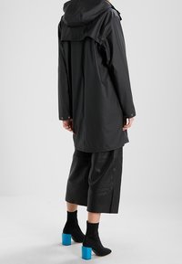 Sort vandtæt frakke med hætte, med en løs pasform og bagventilation. Kombineret med brede læderbukser og sorte ankelstøvler med blå blokhæl.