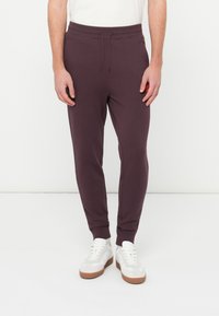 Mężczyzna noszący ciemnofioletowe spodnie dresowe z troczkiem w pasie i białe trampki, stojący na tle jednolitego białego tła.