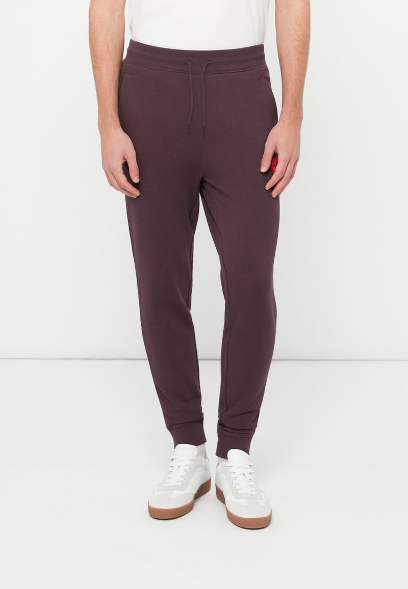 Mężczyzna noszący ciemnofioletowe spodnie dresowe z troczkiem w pasie i białe trampki, stojący na tle jednolitego białego tła.