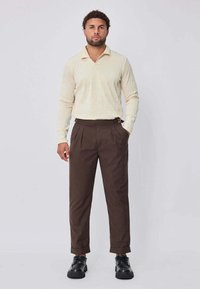 Polo à manches longues jaune clair à rayures, associé à un pantalon plissé marron foncé et des chaussures noires à lacets. Tenue décontractée.