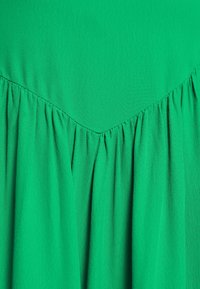 Vihreä mekko, jossa on teksturoitu, kevyt kangas. Ominaisuuksiin kuuluu V-muotoinen halkio ja pystysuorat laskokset, jotka luovat ilmavan muotoilun.