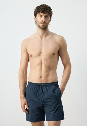 Man with short curly hair wearing navy blue swim shorts, standing with one hand in pocket against plain background.