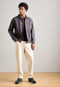 Mann steht mit den Händen in den Taschen, trägt eine dunkelgraue Jacke, ein braunes Hemd, cremefarbene Hosen und schwarze Sneakers auf einem Holzboden.