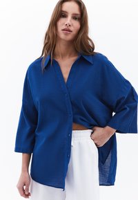Camicia blu a bottone, dal taglio ampio e realizzata in tessuto testurizzato, con colletto classico e maniche a tre quarti, abbinata a pantaloni bianchi.