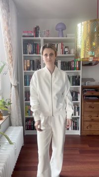 Persona con chaqueta y pantalones blancos de pie frente a una estantería llena de libros, al lado de una ventana con cortinas suaves y plantas de interior.
