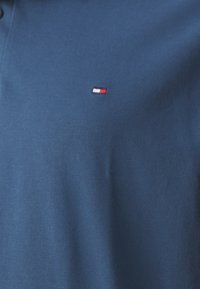 Námorně modré polo tričko vyrobené z texturované látky, s malým vyšívaným logem v červené, bílé a modré barvě na levé hrudi.