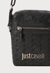 Svart lærveske med preget logo mønster, sølvfarge glidelås og metallbeslag. Har en fremtredende "Just Cavalli"-logo på framsiden.