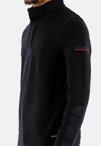Maglione nero a maglia con colletto alto, dotato di zip a metà, dettagli in tessuto blu navy sulle spalle e una striscia rossa sulla manica.