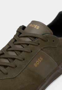 Olivengrønn semsket sneaker med lærakenter, med gull "BOSS"-logo, runde lisseløkker og en teksturert gummisåle.