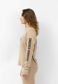 Beige longsleeve top met een ronde hals, met "UNBORED ONBOARD" in zwart op de mouw en een kleurrafische afbeelding op de borst.