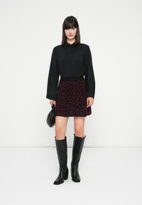 Mujer vistiendo una blusa negra de manga larga, falda de leopardo burdeos, botas negras hasta la rodilla, sosteniendo un pequeño bolso negro contra un fondo blanco.