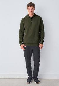Sudadera con capucha de color verde oliva con un patrón texturizado, capucha con cordón y acentos negros, combinada con pantalones negros ajustados y zapatillas deportivas.