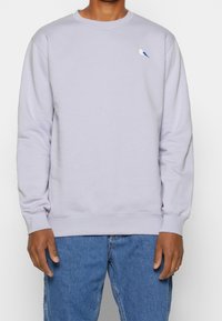 Pullover viola chiaro in cotone, con un piccolo logo ricamato blu e bianco di un uccello sul lato sinistro del petto e polsini a costine.