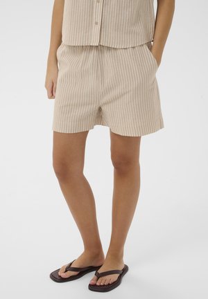 Osoba ubrana w beżowe i białe pionowo paskowane szorty oraz dopasowany top bez rękawów, w ciemnobrązowych klapkach, stojąca na jednolitym tle.