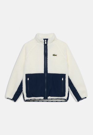Veste polaire blanche et marine à fermeture éclair, avec deux poches avant marine et un petit logo crocodile vert sur la poitrine.