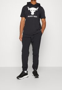 Musta hupullinen t-paita, jossa on valkoinen härän kuva ja teksti "PROJECT ROCK", yhdistettynä mustiin joggereihin ja mustiin urheilukenkiin.