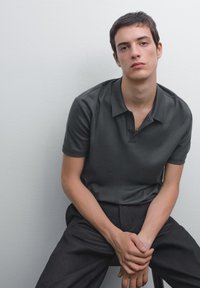 Mörkgrå polo skjorta med en mjuk textur, med en knappad krage och korta ärmar, tillsammans med mörka byxor. Modell som sitter mot en neutral bakgrund.