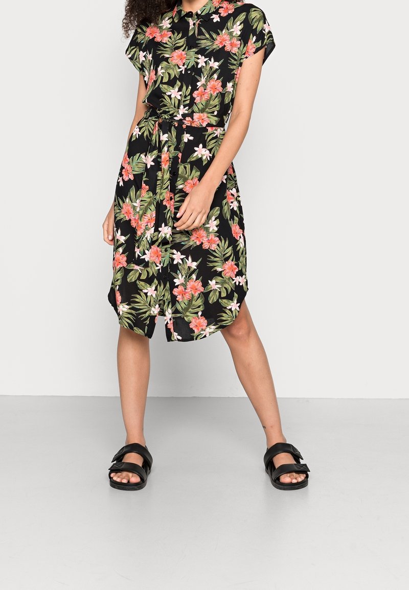 Femme portant une robe noire à manches courtes, longueur genoux, avec un imprimé floral rose et blanc et des sandales noires à brides.