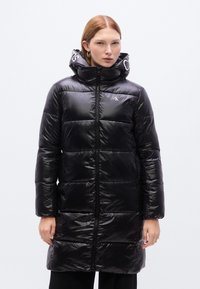 Μαύρο φουσκωτό παλτό με γυαλιστερό φινίρισμα, πλήρες φερμουάρ, ψηλό γιακά και λογότυπο στη κουκούλα. Διαθέτει κάθετο καπιτονέ σχέδιο και πλαϊνές τσέπες.