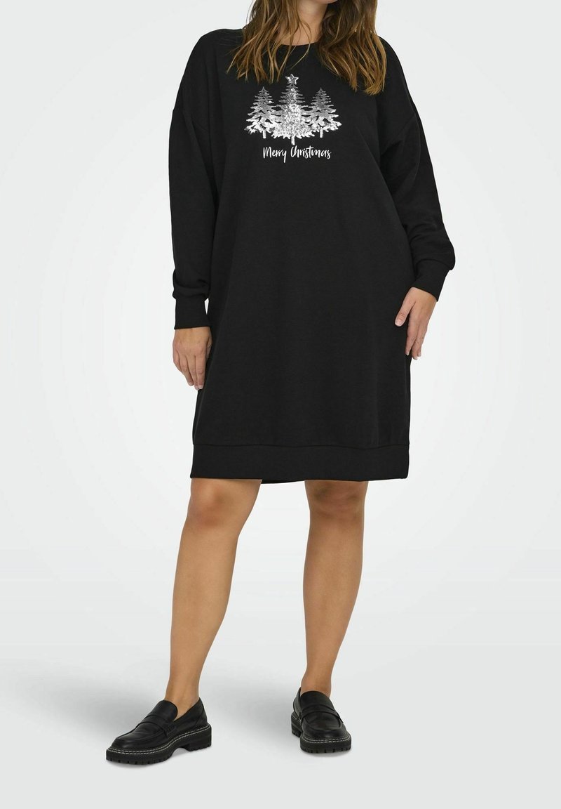 Femme portant une robe noire ample à manches longues avec un motif sapin de Noël argenté et le texte « Joyeux Noël », associée à des mocassins noirs.