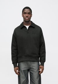 Camisola preta com gola, pala com quatro botões e emblema no peito esquerdo. O tecido parece macio e ligeiramente texturizado.