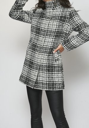 Femme portant un manteau à manches longues à carreaux noir et blanc avec un col montant, associé à un pantalon en cuir noir, sur un fond uni.