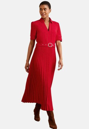 Woman wearing a red pleated midi dress with short sleeves, collar, belt at waist, and brown suede boots, walking against white background.