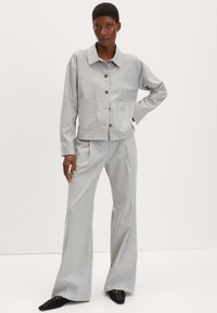Veste ajustée gris clair avec deux poches avant et fermeture à boutons, associée à un pantalon large assorti. Modèle portant des chaussures noires à bout pointu.