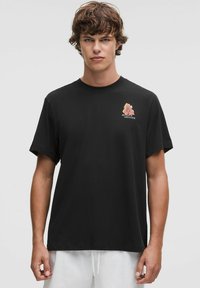 Tricou negru din bumbac, cu mâneci scurte, având o grafică florală colorată și text în zona superioară stângă a pieptului, cu un design lejer.