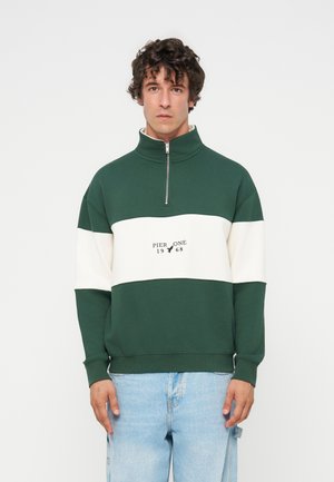 Jeune homme aux cheveux bouclés portant un pull vert et blanc à fermeture éclair quart de zip avec le texte "Pier One 1968", associé à un jean bleu clair.
