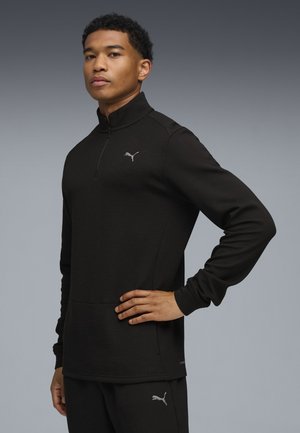 Jonge man staat met zijn hand op zijn heup, draagt een zwarte Puma lange mouwen zip-up top en bijpassende zwarte broek tegen een neutrale achtergrond.