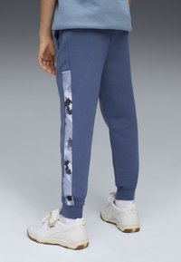 Joggers de algodón azules con un panel abstracto gris texturizado en el lateral. Puños acanalados y bolsillos. Llevados con zapatillas blancas.