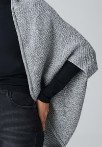 Nahaufnahme einer Person, die ein schwarzes Langarmoberteil und dunkle Jeans trägt, mit einem strukturierten grauen Strickschal über einem Arm drapiert.