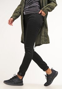 Un jean skinny noir assorti à un t-shirt gris foncé et un long manteau vert olive. Des chaussures de sport noires avec un matériau texturé et des lacets.